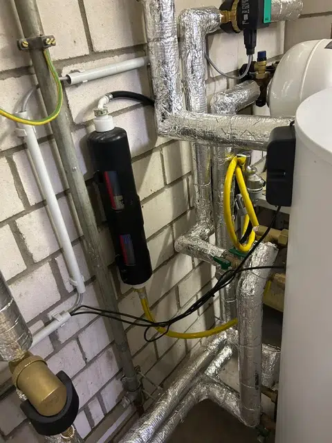 Schwarzer Schlammabscheider an der Wand, gelbe Gasleitung, gedämmte Heizungsrohre und Warmwasserspeicher im modernisierten Heizungsraum